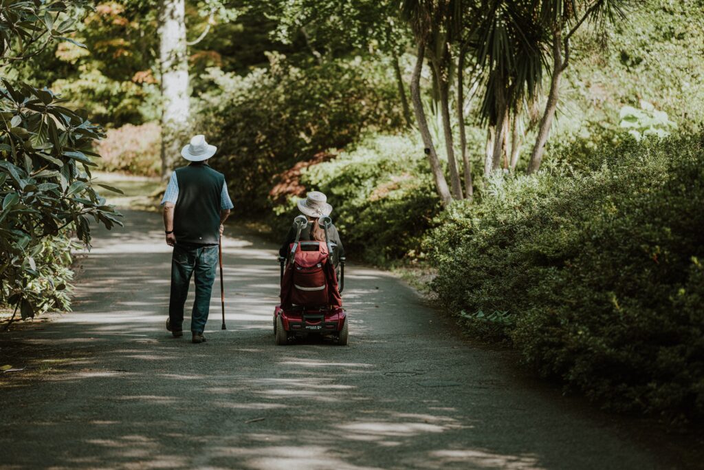 One person using a walking stick and another using a mobility scooter walk side-by-side. 