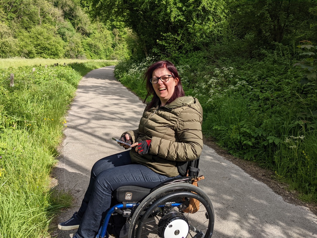 A photo of Naomi, on a road in the countryside, sitting in a blue wheelchair with grey wheels and a black seat cushion. Naomi is wearing a green coat and making an odd face.